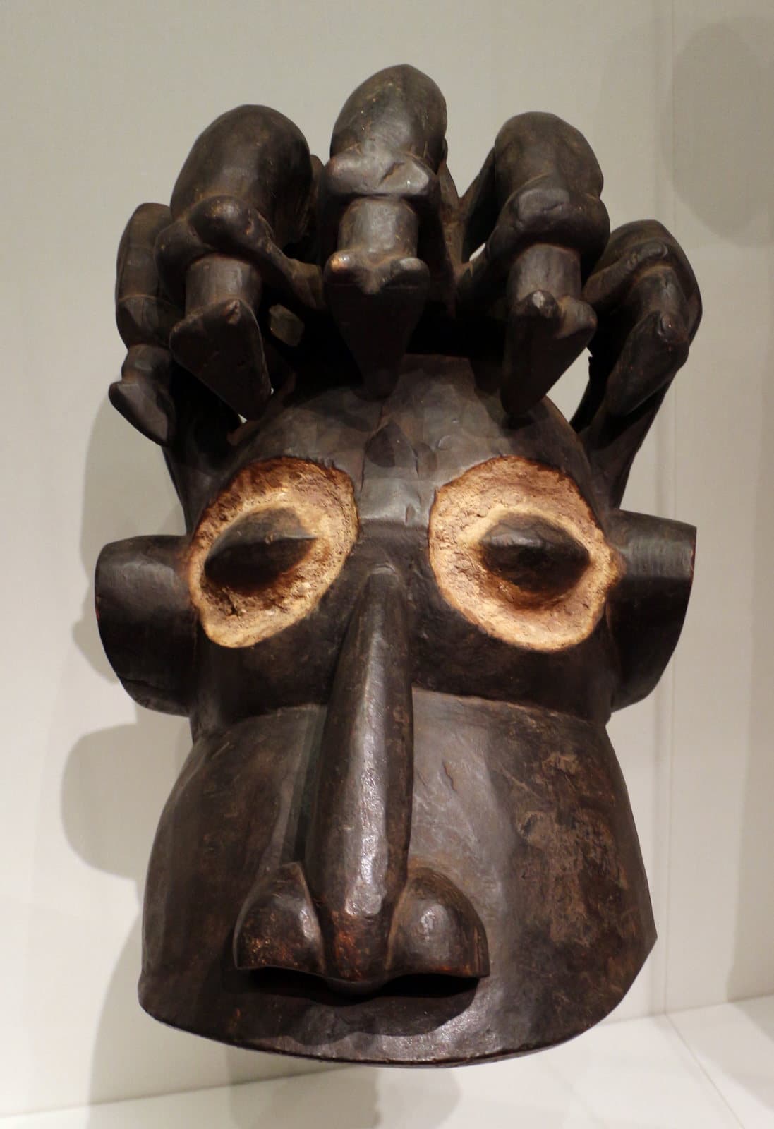 Masque-heaume en bois sombre du peuple Kom, aux yeux cerclés de blanc, surmonté d'une rangée de petites figures sculptées.