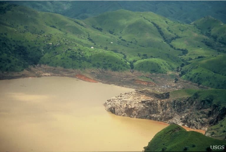 Vue aérienne du lac Nyos aux eaux troubles et rougeâtres après l'éruption limnique de 1986, falaise rocheuse au premier plan, collines vertes du Cameroun en arrière-plan.