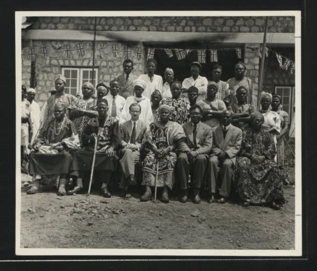 Photographie noir et blanc de groupe : des notables camerounais en tenue traditionnelle et des fonctionnaires en costume, assis et debout devant un bâtiment colonial décoré de drapeaux britanniques, à Bamenda en 1958.