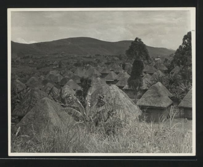 Photographie noir et blanc en plongée d'un village aux toits de chaume coniques serrés les uns contre les autres, entouré de collines herbeuses.