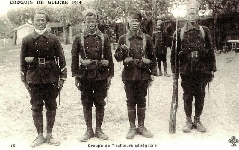 Quatre soldats tirailleurs sénégalais en uniforme sombre, chéchia claire, posant en groupe devant des cases, vers 1914-1918.