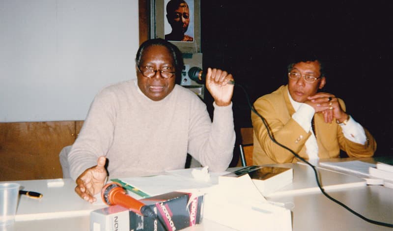Photographie couleur de deux hommes assis à une table avec des microphones ; à droite, le professeur Théophile Obenga, en 2008.