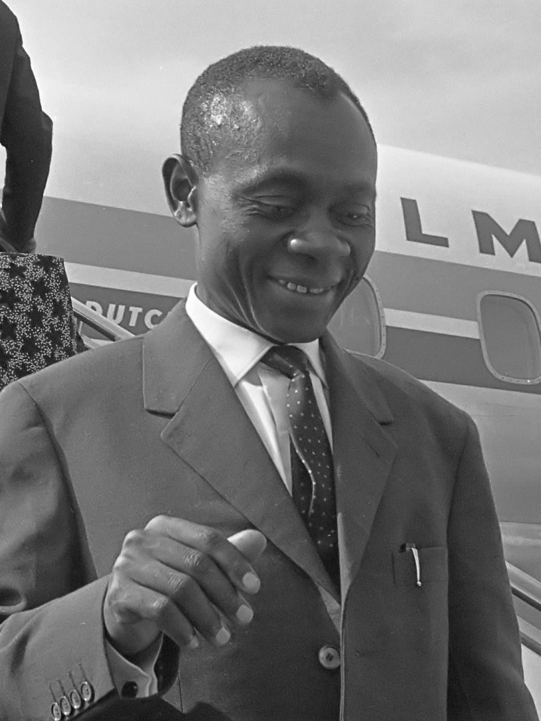 Photographie en noir et blanc d'un homme souriant en costume sombre et cravate, descendant la passerelle d'un avion, 1964.