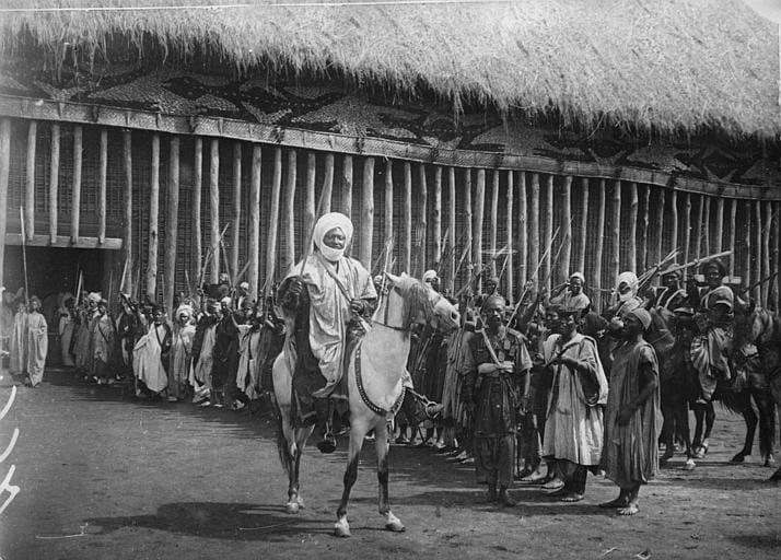 Photographie en noir et blanc de 1917 : un homme en turban blanc monté à cheval sort d'une longue bâtisse à toit de chaume, encadré par une file de gardes en tenue traditionnelle portant des lances et des fusils.