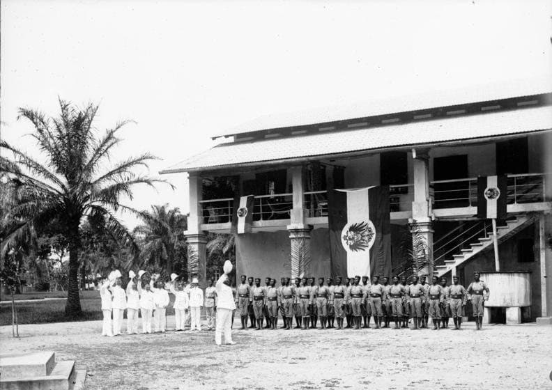 Photographie ancienne en noir et blanc d'une troupe de policiers africains en uniforme alignés devant un bâtiment colonial, à Douala en 1901.