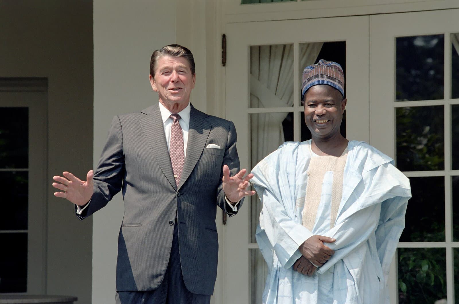 Photo couleur de Ronald Reagan, en costume gris, bras ouverts, et Ahmadou Ahidjo, en boubou clair et bonnet brodé, tous deux souriants, devant la roseraie de la Maison-Blanche.