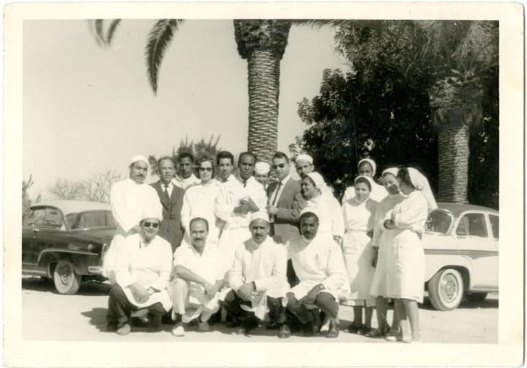 Photographie noir et blanc d'une quinzaine de médecins, infirmières et aides-soignants en blouses blanches, posant en groupe devant des palmiers et deux automobiles anciennes.