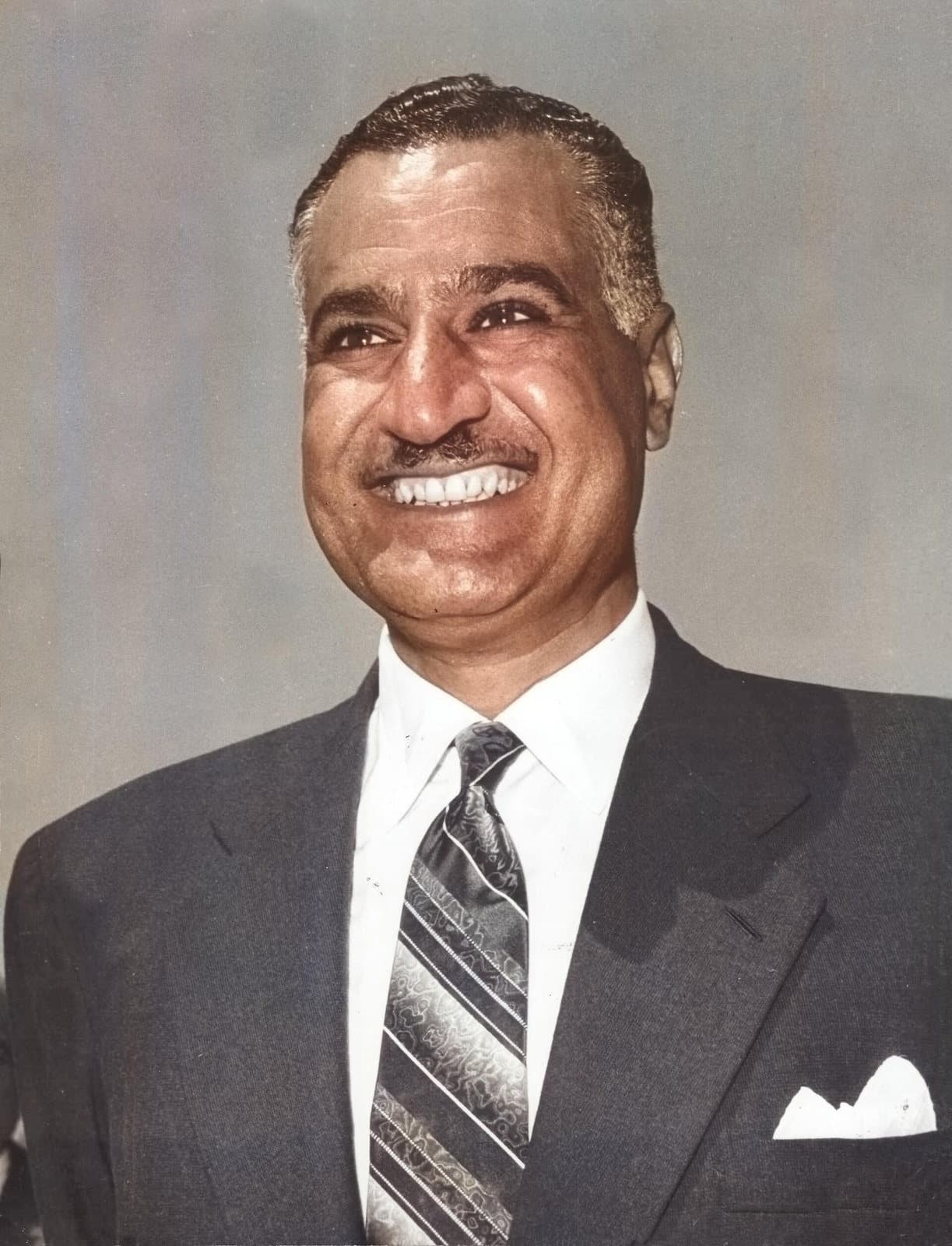 Portrait en couleur du président égyptien Gamal Abdel Nasser, souriant, en costume sombre et cravate rayée, sur fond gris uni.