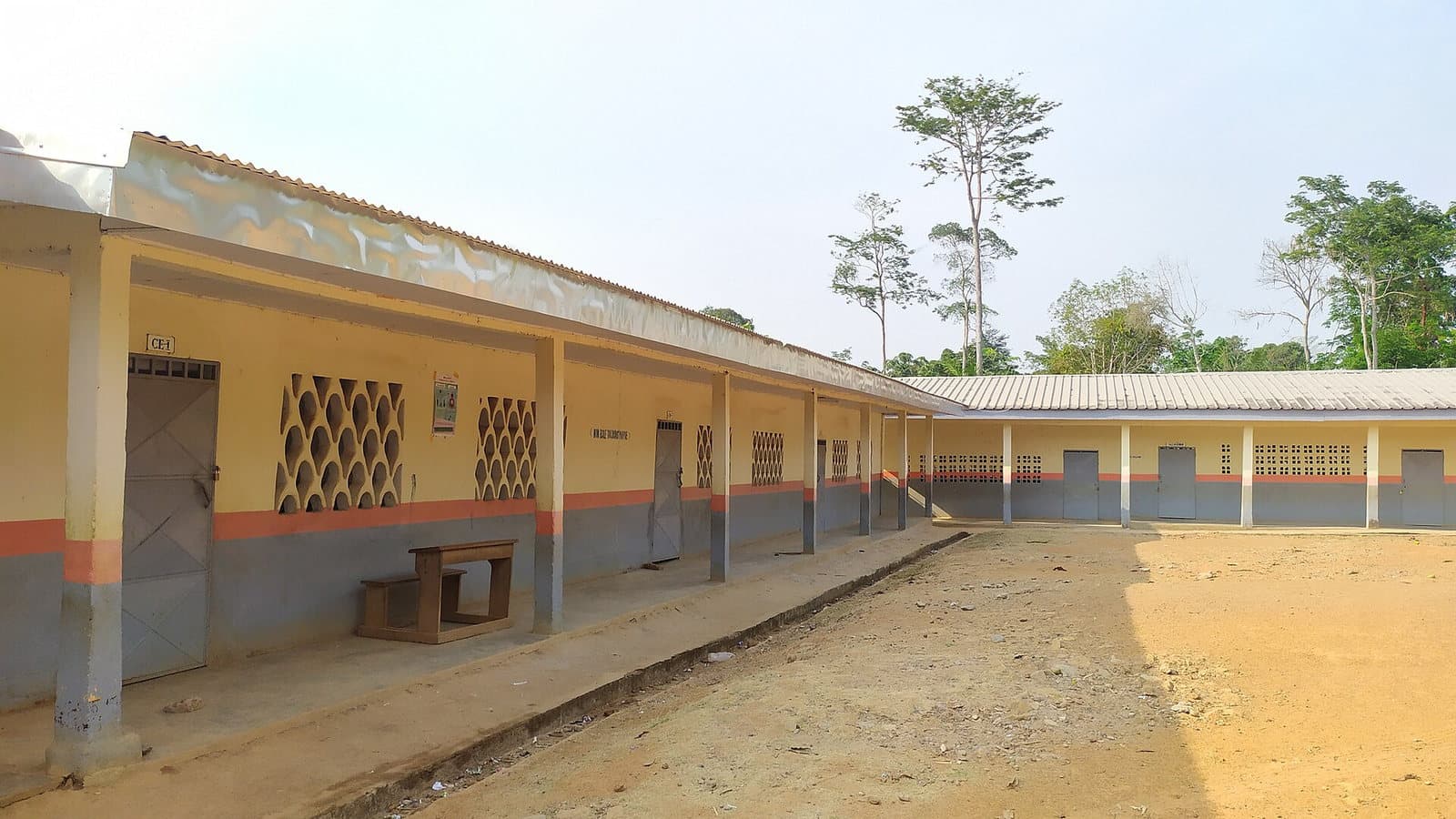 Vue extérieure d'un long bâtiment scolaire aux murs jaune et gris sous un toit de tôle, en bordure d'une cour de terre rouge, avec la forêt en arrière-plan.