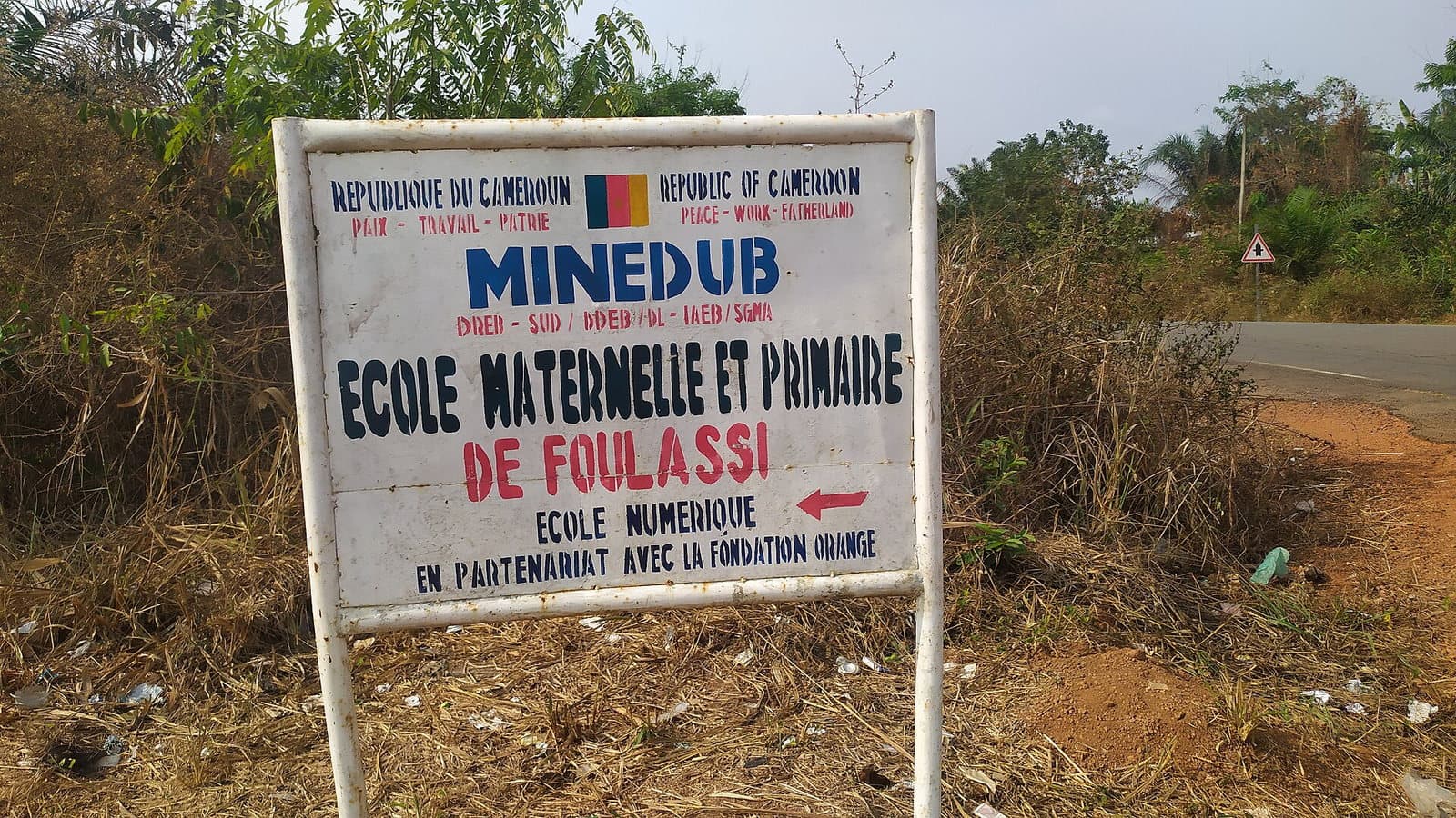 Pancarte métallique en bordure de route, en français et en anglais : « République du Cameroun, MINEDUB, École maternelle et primaire de Foulassi », plantée au milieu de hautes herbes.