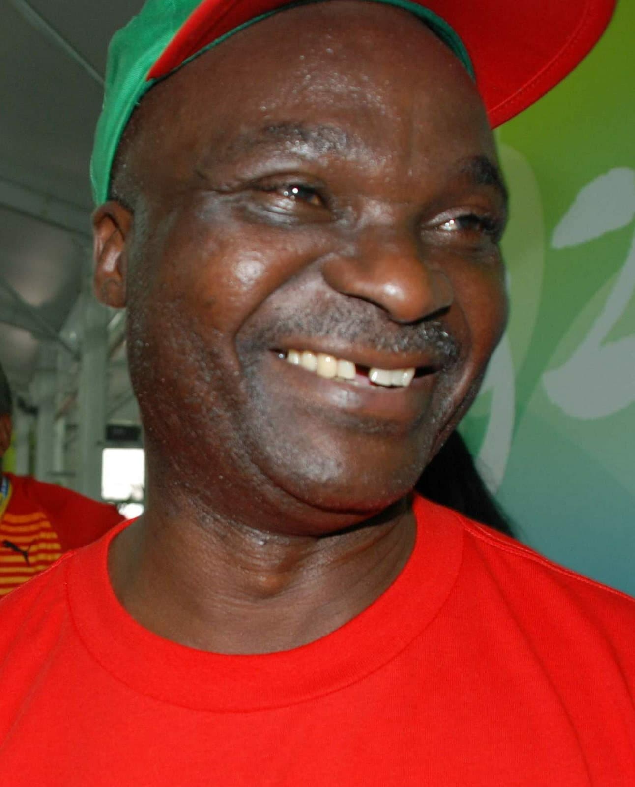 Portrait souriant de Roger Milla portant une casquette et un maillot aux couleurs du Cameroun.