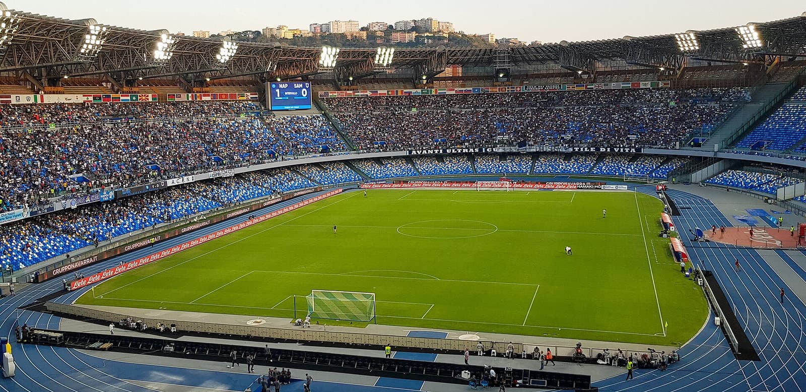 Vue plongeante sur un stade de football napolitain plein, projecteurs allumés, tableau d'affichage visible.