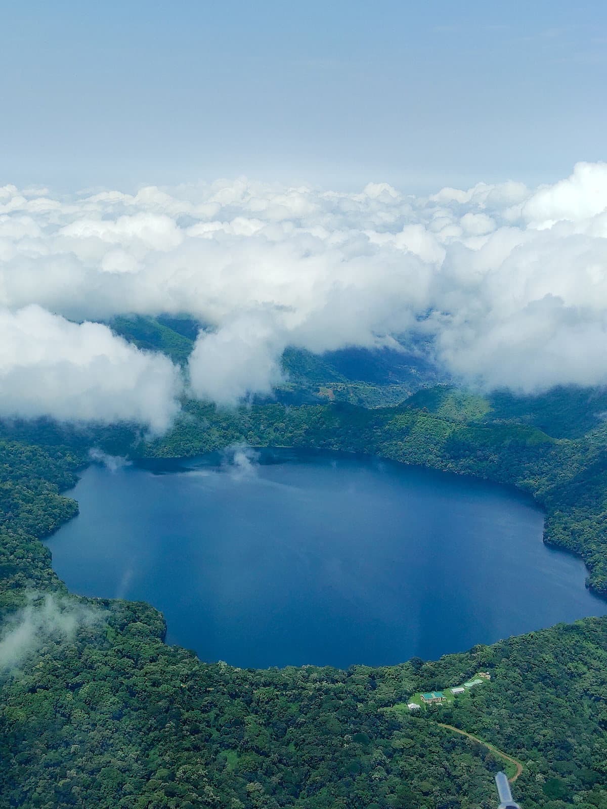Vue aérienne d'un lac de cratère bleu entouré de forêt de montagne et de nuages, au Cameroun.