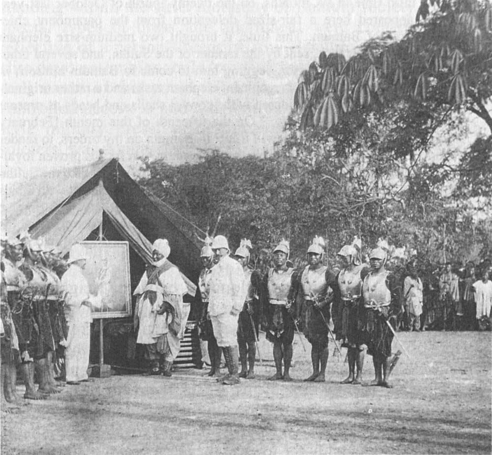 Photographie ancienne : un officier en tenue blanche présente un tableau encadré à un dignitaire africain devant une tente de campagne, entouré de gardes en armure, 1906.