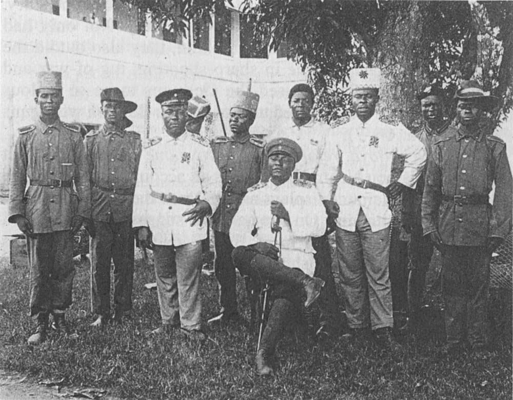 Photographie ancienne : un groupe d'hommes en uniforme militaire posent devant un bâtiment colonial à Douala, un homme assis au centre, janvier 1908.
