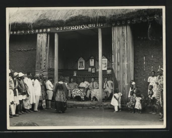 Photographie ancienne en noir et blanc : dans la cour intérieure d'un palais traditionnel des Grassfields, au toit de chaume et aux piliers ornés de motifs sculptés, une assemblée d'hommes se tient devant des dignitaires assis.