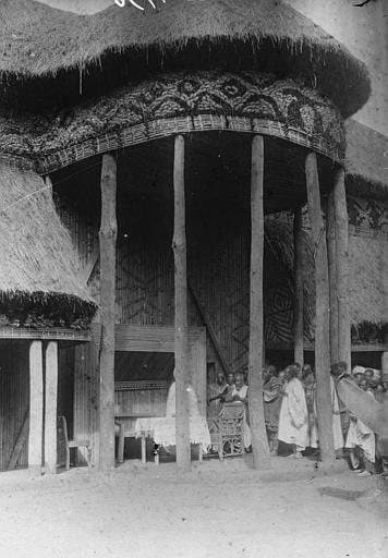 Photographie en noir et blanc de 1917 : sous un grand pavillon de chaume à piliers de bois sculptés ornés de motifs géométriques, un groupe d'hommes en tenue traditionnelle se tient debout et assis autour d'une table, dans le palais du sultan de Foumban.