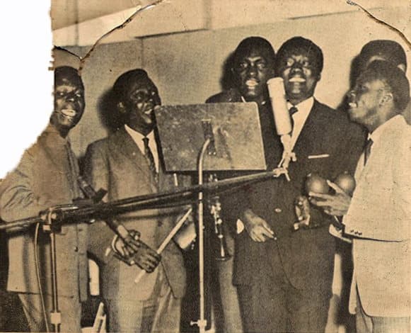 Photographie noir et blanc de l'orchestre congolais African Jazz en studio d'enregistrement en 1961, autour d'un microphone.