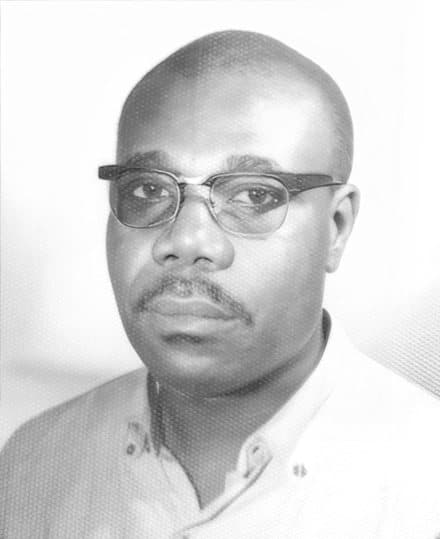 Photo d'identité en noir et blanc de Manu Dibango en 1964, crâne rasé, lunettes à monture noire, chemise claire à col ouvert.