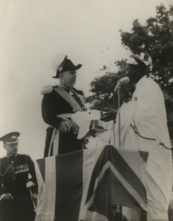 Photographie noir et blanc : un gouverneur britannique en tenue de cérémonie remet un paquet enveloppé à un chef africain en robe blanche coiffé d'un turban, devant un pavillon orné de l'Union Jack.