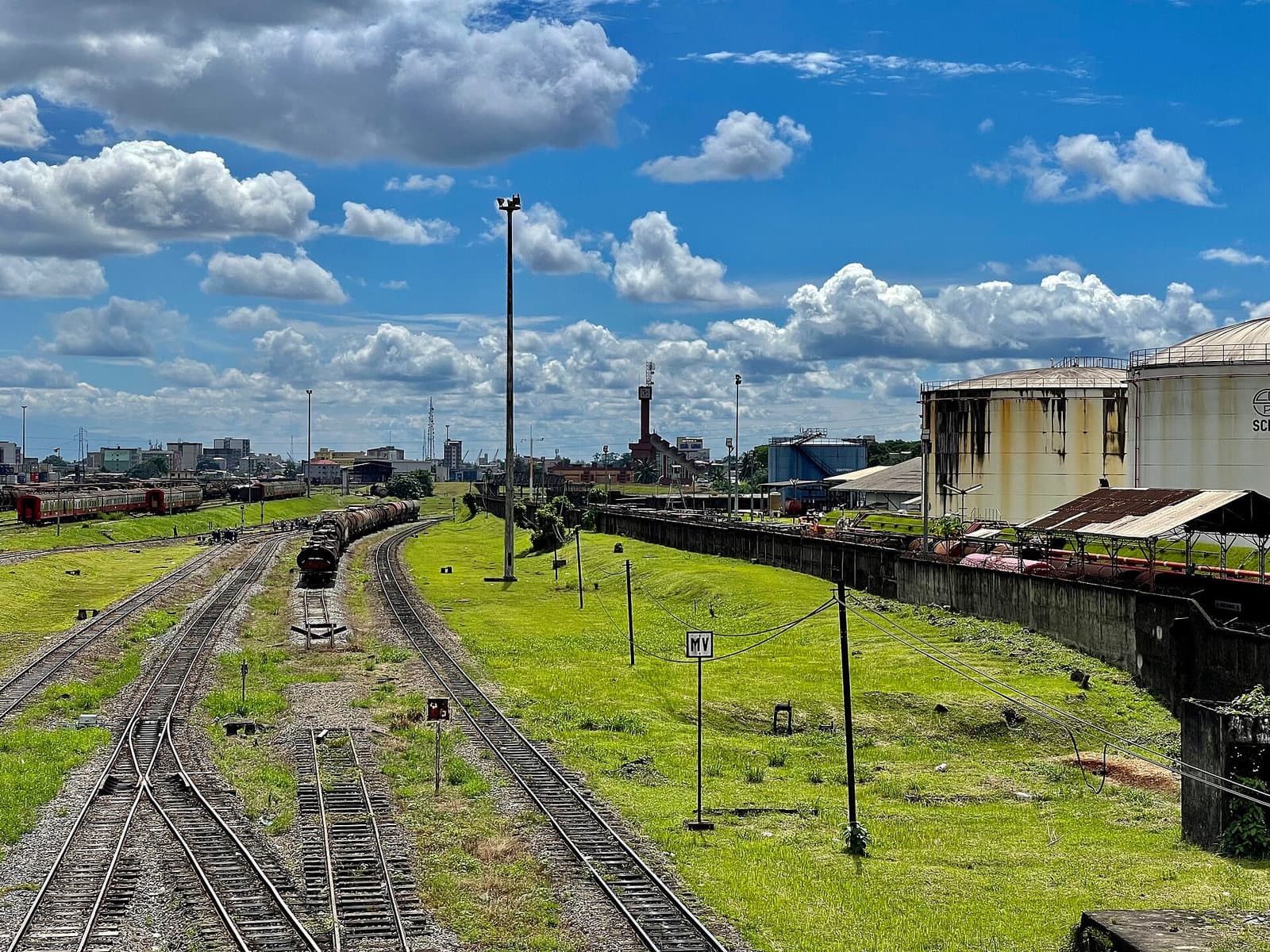 Grands réservoirs cylindriques de stockage pétrolier rouillés au sommet, voies ferrées et bâtiments industriels d'un dépôt de la SCDP à Douala, sous un ciel bleu à nuages.