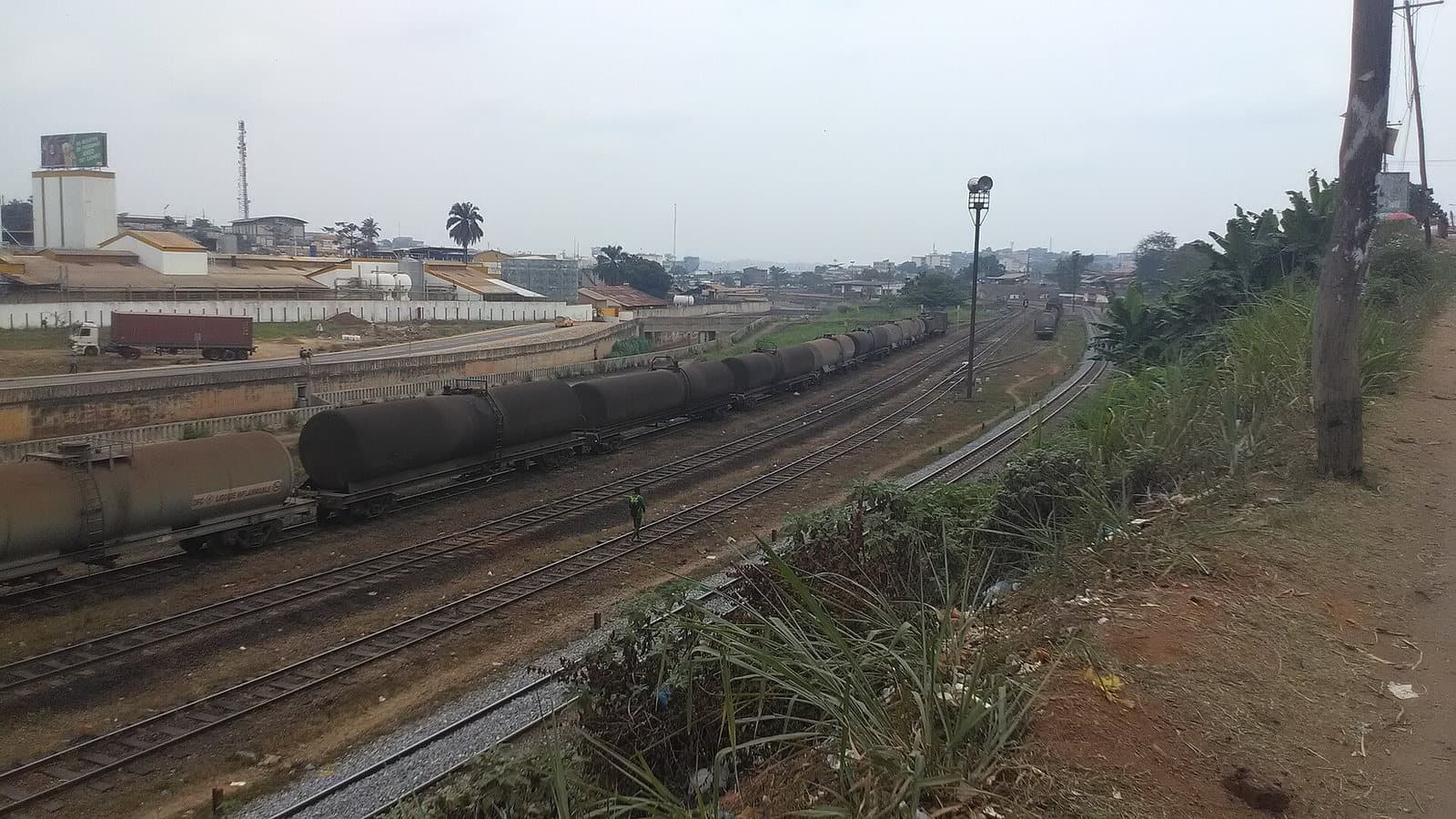 Voies ferrées industrielles à Nsam, Yaoundé, avec des wagons-citernes garés en enfilade, un bâtiment industriel et des collines urbaines à l'arrière-plan.