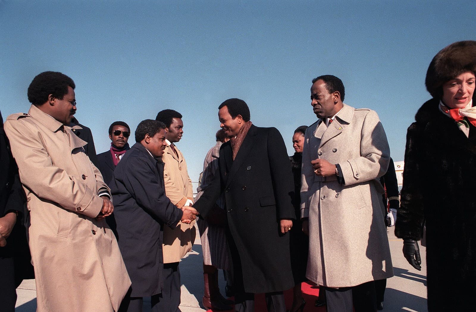 Photographie couleur de Paul Biya en manteau sombre serrant la main d'un dignitaire, entouré d'une délégation en manteaux d'hiver sur le tarmac d'une base aérienne américaine.