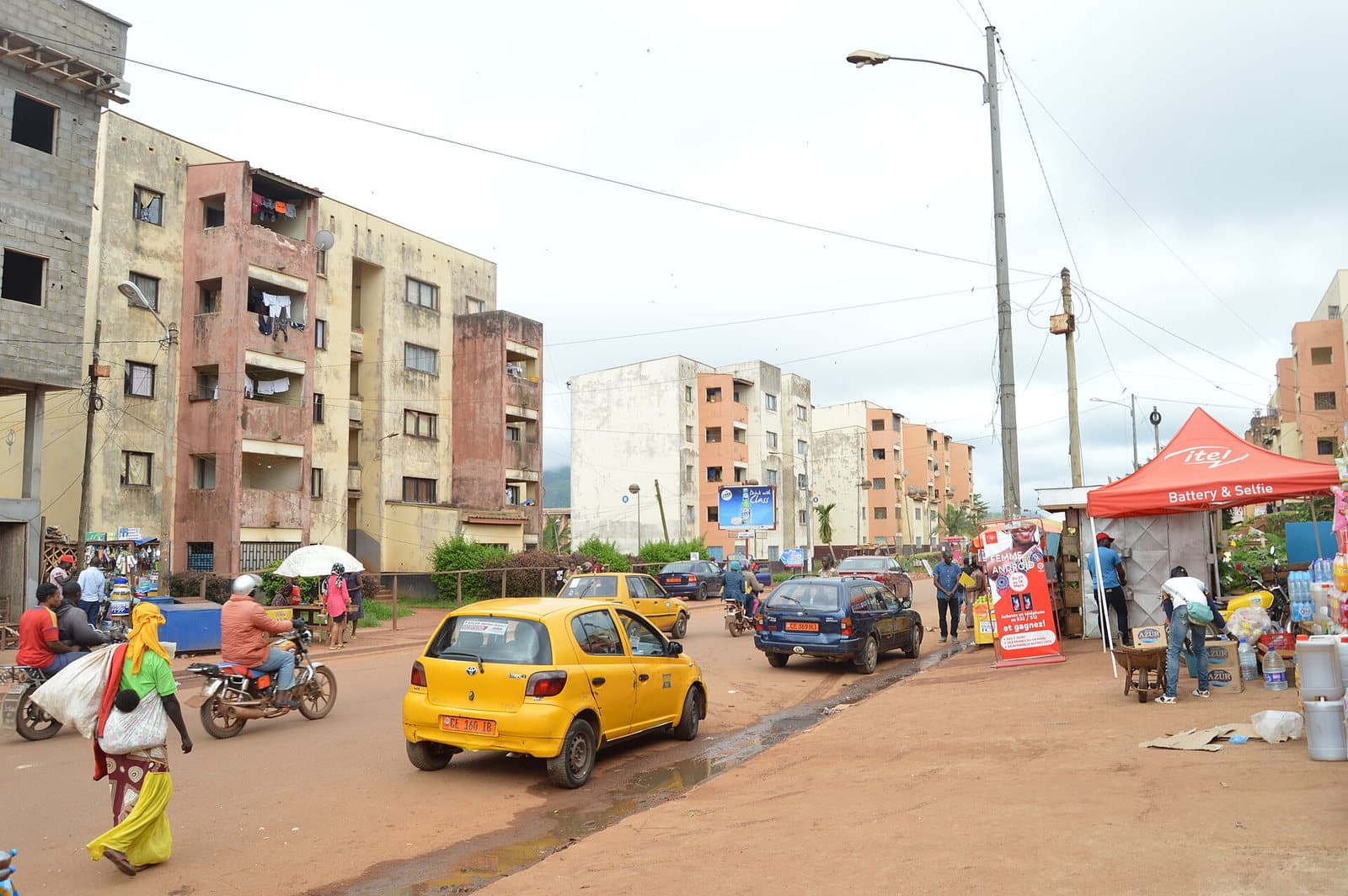 Rue animée du quartier de Mendong à Yaoundé, avec des immeubles résidentiels décrépis, un taxi jaune, des motos-taxis, un marché de rue et des passants.