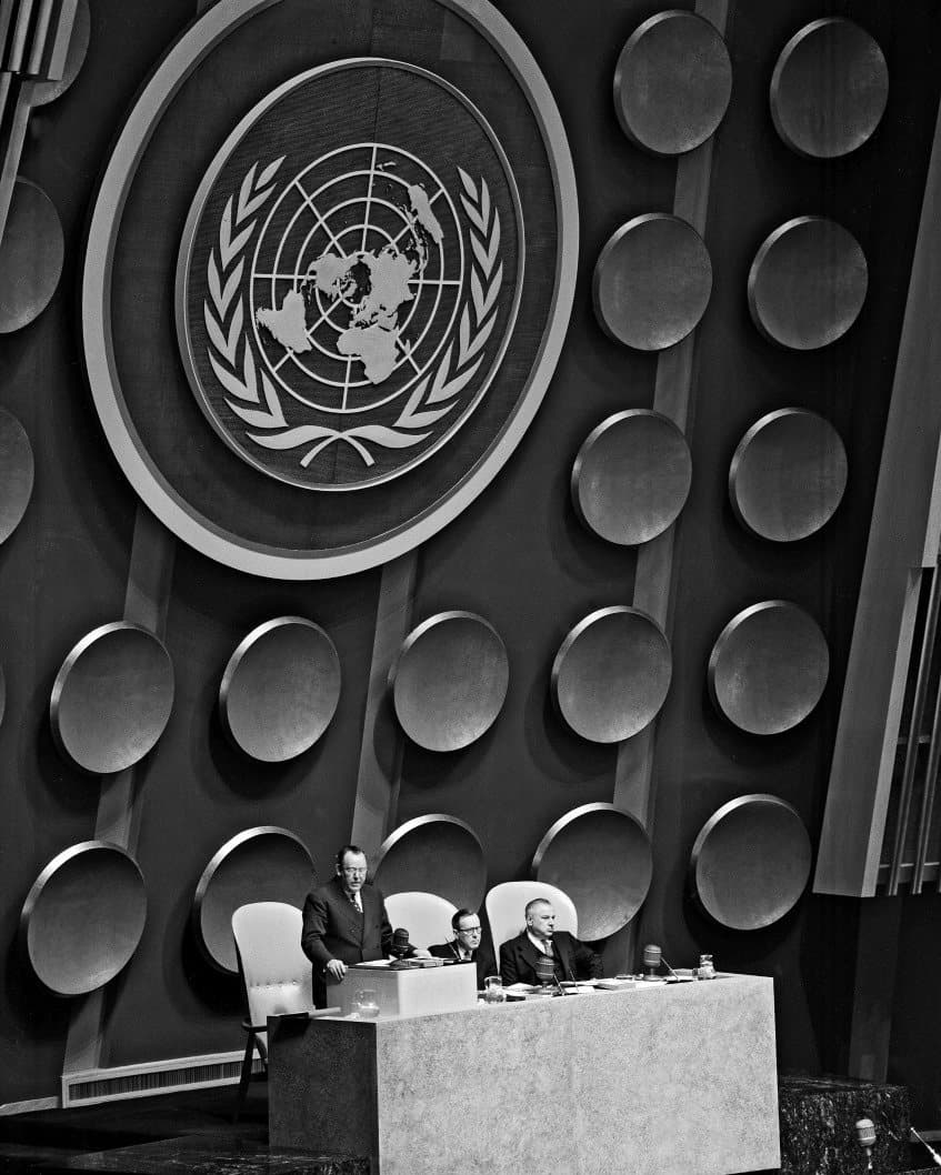 Estrade de l'Assemblée générale de l'ONU à New York en 1952, sous l'emblème des Nations unies, avec trois hommes assis et debout au pupitre